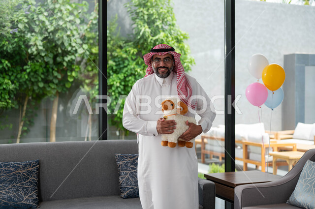 ايماءات تدل على الفرح و السعادة, رجل كبير السن عربي خليجي سعودي يرتدي الزي السعودي التقليدي و يحمل بيده دمية على شكل خاروف عيد الاضحى المبارك, أعياد ومناسبات اسلامية, الاحتفال بالعيد السعيد .