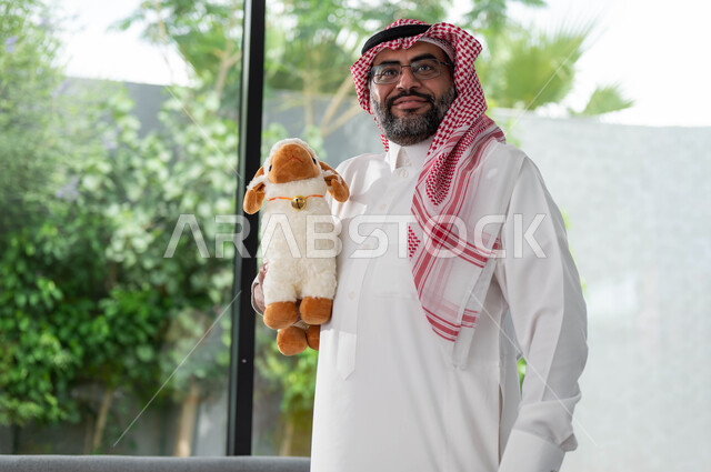 رجل كبير السن عربي خليجي سعودي يرتدي الزي السعودي التقليدي و يحمل بيده دمية على شكل خاروف عيد الاضحى المبارك, أعياد ومناسبات اسلامية, ايماءات تدل على السعادة, الاحتفال بالعيد السعيد .