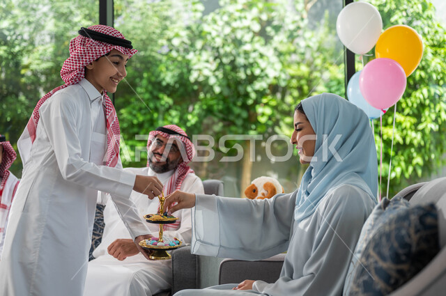 Congratulating on the advent of Eid, presenting Eid sweets and cakes, welcoming and honoring guests, a Saudi Arabian Gulf family celebrating Eid, gestures indicating joy and happiness, family visits and gatherings, Islamic holidays and happy occasions.