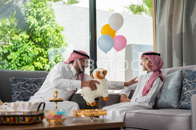 التسامر والضحك مع الاحفاد في عيد الاضحى المبارك ،رجل كبير السن عربي خليجي سعودي يجلس في غرفة المعيشة يرتدي الزي التقليدي السعودي يقوم باللعب مع حفيده ، انشطه ترفيهيه في الاعياد والمناسبات العائليه, ايماءات تدل على السعادة