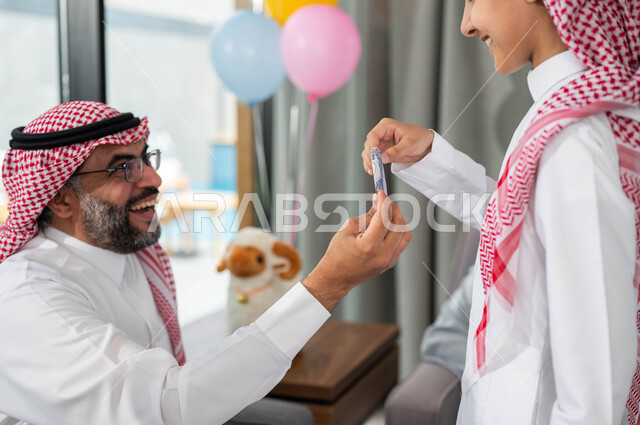تقديم العيدية للأحفاد, أعياد ومناسبات اسلامية, أجواء العيد في المجتمع العربي السعودي,  التجمعات والزيارات الاسرية في عيد الاضحى المبارك, ايماءات تدل على السعاده , رجل مسن عربي خليجي سعودي يحتفل في العيد مع الابناء في ايام العيد السعيد في السعوديه