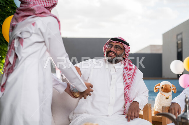 قضاء اوقات ممتعة مع  الابناء في الشاليهات والمنتجعات في ايام العيد السعيد في السعوديه, فتى عربي خليجي سعودي يرتدي الزي التقليدي السعودي ويقوم بتقديم توزيعات عيد الاضحى المبارك,  التجمعات والزيارات الاسرية, أعياد ومناسبات اسلامية, ايماءات تدل على السعاده 