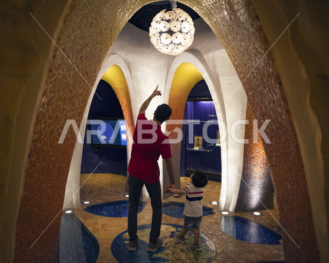 رجل خليجي عربي سعودي برفقة ابنه داخل مركز الملك عبد العزيز الثقافي العالمي في مدينة الظهران في المملكة العربية السعودية ، أماكن سياحية و منارة ثقافية ومعالم مشهورة في السعودية ، الديكور و الفن والتصميم الهندسي المعماري الحديث.