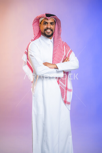 إيماءات وجه مبتسمة ويدين مكتفة، بورتريه لشاب وسيم عربي خليجي يرتدي الثوب و الشماغ السعودي التقليدي وينظر إلى الكاميرا، خلفية ملونة.