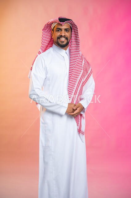Portrait of a handsome young man wearing the traditional Saudi thawb and shemagh, standing straight and looking at the camera, face gestures and a smile of happiness and pleasure, colored background.