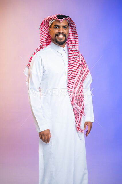 Portrait of a handsome young man wearing the traditional Saudi thawb and shemagh, standing straight and looking at the camera, face gestures and a smile of happiness and pleasure, colored background.