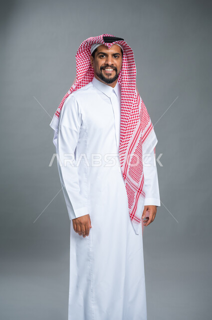 بورتريه شاب وسيم يرتدي الثوب و الشماغ السعودي التقليدي يقف باستقامة وينظر نحو الكاميرا, ايماءات وجه و ابتسامة سعادة و سرور, خلفية رمادية.