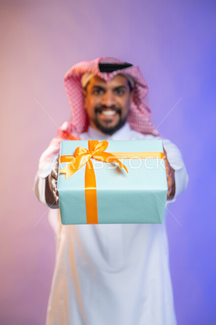 إيماءات وجه ويدين تدل على الفرح والسعادة بالهدايا و التهنئات، بورتريه لشاب عربي سعودي خليجي يرتدي الثوب و الشماغ السعودي التقليدي يحمل بيده علبة هدايا،  أعياد ومناسبات، مفاجأت سعيده, خلفية ملونة.