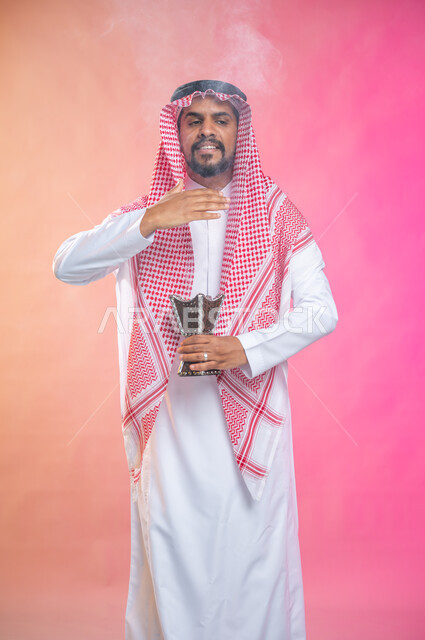 التطيب بالبخور والعود، بورتريه لرجل عربي سعودي خليجي يرتدي الثوب و الشماغ السعودي التقليدي يتعطربحامل البخور ( المبخرة ) ، استخدام خشب العود في المناسبات والأعياد, العادات والتقاليد السعودية, خلفية ملونة.