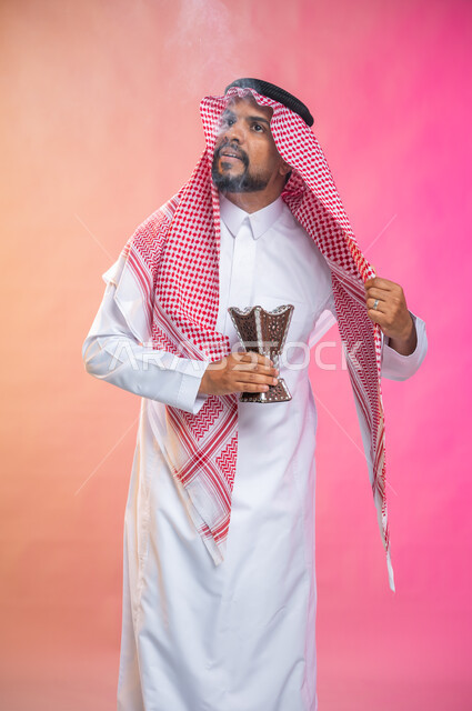 التطيب بالبخور والعود، بورتريه لرجل عربي سعودي خليجي يرتدي الثوب و الشماغ السعودي التقليدي يتعطربحامل البخور ( المبخرة ) ، استخدام خشب العود في المناسبات والأعياد, العادات والتقاليد السعودية, خلفية ملونة.