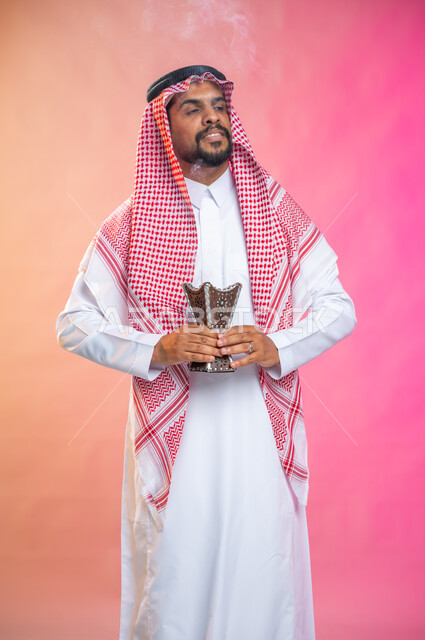 التطيب بالبخور والعود، بورتريه لرجل عربي سعودي خليجي يرتدي الثوب و الشماغ السعودي التقليدي يتعطربحامل البخور ( المبخرة ) ، استخدام خشب العود في المناسبات والأعياد, العادات والتقاليد السعودية, خلفية ملونة.