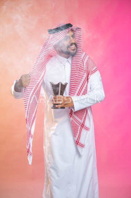 استخدام خشب العود في المناسبات والأعياد, التطيب بالبخور والعود، بورتريه لرجل عربي سعودي خليجي يرتدي الثوب و الشماغ السعودي التقليدي يتعطربحامل البخور ( المبخرة ) ، العادات والتقاليد السعودية, خلفية ملونة.