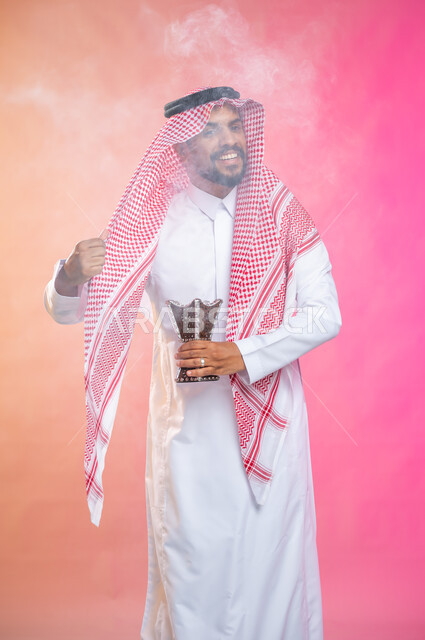 استخدام خشب العود في المناسبات والأعياد, التطيب بالبخور والعود، بورتريه لرجل عربي سعودي خليجي يرتدي الثوب و الشماغ السعودي التقليدي يتعطربحامل البخور ( المبخرة ) ، العادات والتقاليد السعودية, خلفية ملونة.