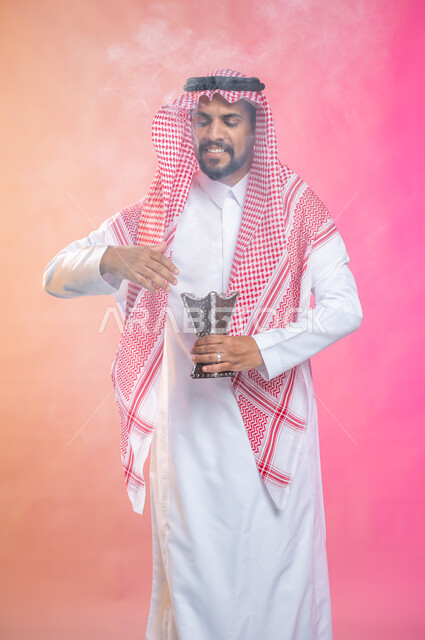 استخدام خشب العود في المناسبات والأعياد, التطيب بالبخور والعود، بورتريه لرجل عربي سعودي خليجي يرتدي الثوب و الشماغ السعودي التقليدي يتعطربحامل البخور ( المبخرة ) ، العادات والتقاليد السعودية, خلفية ملونة.