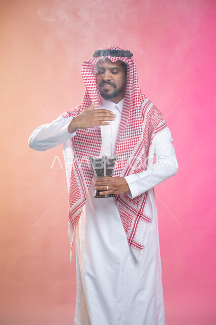 استخدام خشب العود في المناسبات والأعياد, التطيب بالبخور والعود، بورتريه لرجل عربي سعودي خليجي يرتدي الثوب و الشماغ السعودي التقليدي يتعطربحامل البخور ( المبخرة ) ، العادات والتقاليد السعودية, خلفية ملونة.