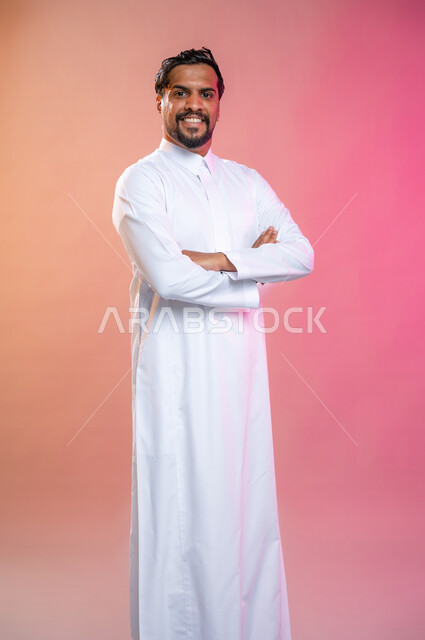 إيماءات وجه مبتسمة ويدين مكتفة، بورتريه لشاب وسيم  عربي خليجي يرتدي الثوب السعودي التقليدي وينظر إلى الكاميرا،  خلفية ملونة.
