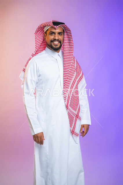 Portrait of a handsome young man wearing the traditional Saudi thawb and shemagh, standing straight and looking at the camera, face gestures and a smile of happiness and pleasure, colored background.