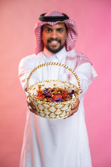 إيماءات وجه تدل على البشاشه والترحيب بالضيوف, بورتريه لرجل عربي سعودي خليجي يرتدي الثوب السعودي التقليدي يحمل بيده سلة من القش مليئه بالحلوى و السكاكر , حسن الضيافه في الأعياد و المناسبات , خلفيه ملونة.