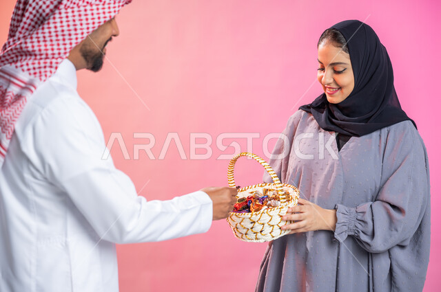 بورتريه لإمرأة محجبة  تحمل سلة قش  وتقدم الحلوى و السكاكر لرجل يرتدي الثوب و الشماغ السعودي التقليدي بمناسبة الأعياد السعيدة ، حسن الضيافة في المناسبات و الأعياد الاسلامية، ايماءات فرحة و سرور و سعادة بالاحتفاليات ، خلفية ملونة.