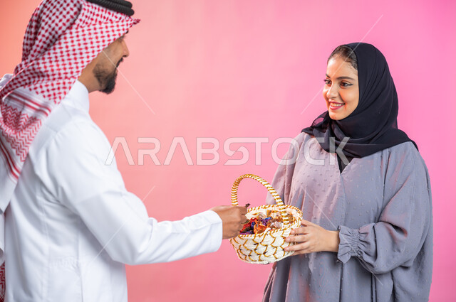 بورتريه لإمرأة محجبة  تحمل سلة قش  وتقدم الحلوى و السكاكر لرجل يرتدي الثوب و الشماغ السعودي التقليدي بمناسبة الأعياد السعيدة ، حسن الضيافة في المناسبات و الأعياد الاسلامية، ايماءات فرحة و سرور و سعادة بالاحتفاليات ، خلفية ملونة.