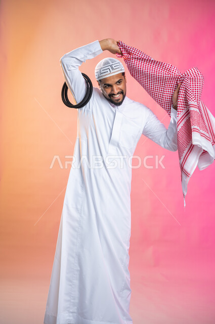 Steps to wear the traditional Saudi shemagh,  Getting ready to go out, adorning, beautifying and arranging, a young Saudi Arab Gulf man wearing a red dress and shemagh, taking care of the external appearance, elegance and elegance, a colored background.