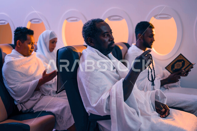 مجموعة من حجاج الخارج يجلسون في مقصورة الطائرة ويرتدون ملابس الاحرام، ايماءات العبادة والتقرب الى الله، قراءة المصحف الشريف، موسم الحج 2023، السفر الى مكة لآداء مناسك الحج والعمرة، فضل العمرة في شهر رمضان، استقبال الحجاج من خارج السعودية