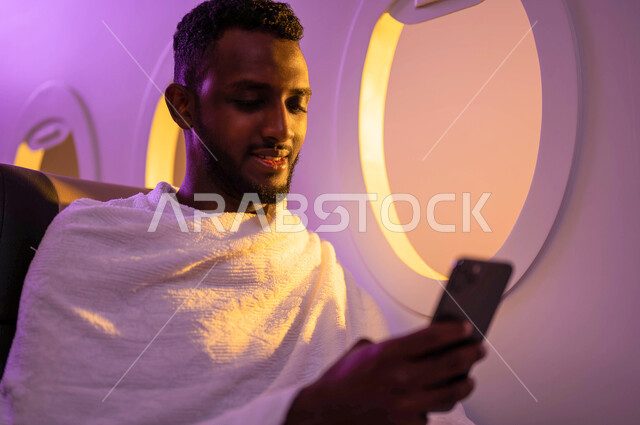 الحجز الالكتروني لحملات الحج والعمرة باستخدام الجوال، السفر الى مكة لآداء مناسك الحج والعمرة، ارتداء ملابس الاحرام، رجل عربي سوداني يستخدم الهاتف المحمول ويجلس في الطائرة، موسم الحج 2023، فضل العمرة في شهر رمضان، خطوط الطيران السعودي