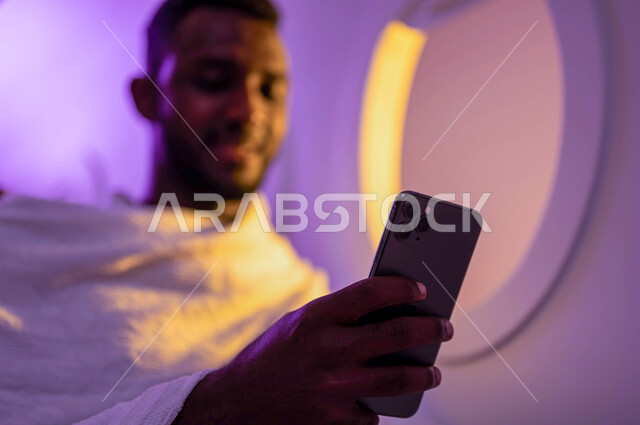 السفر الى مكة لآداء مناسك الحج والعمرة، الحجز الالكتروني لحملات الحج والعمرة باستخدام الجوال، ارتداء ملابس الاحرام، رجل عربي سوداني يستخدم الهاتف المحمول ويجلس في الطائرة، موسم الحج 2023، فضل العمرة في شهر رمضان، خطوط الطيران السعودي