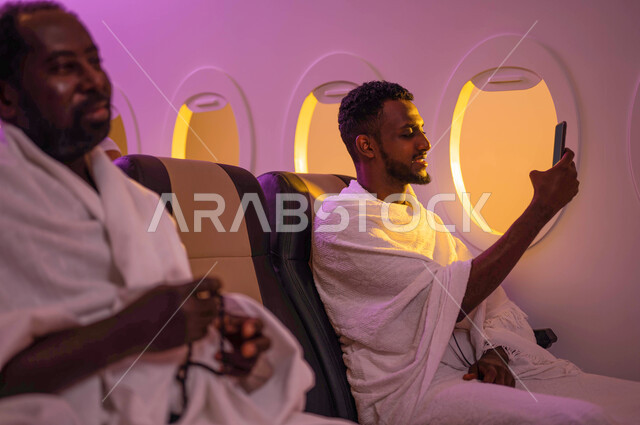 التقاط صور تذكارية من خلال الهاتف المحمول، بث فيديو مباشر، رجلان عربيان من السودان يجلسان في الطائرة ويرتديان ملابس الاحرام، السفر الى مكة لآداء مناسك الحج والعمرة، موسم الحج 2023، آداء العمرة في رمضان، خطوط الطيران السعودي