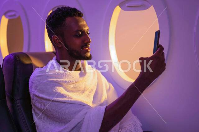 Live video broadcast, taking souvenir photos, a Sudanese Arab man sitting on the plane and using a mobile phone, wearing Ihram clothes, traveling to Mecca to perform the rituals of Hajj and Umrah, the Hajj season 2023, the virtue of Umrah in the month of 