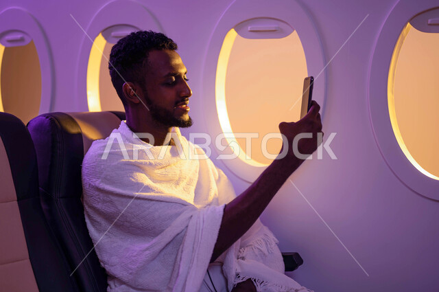 ارتداء ملابس الاحرام، بث فيديو مباشر، التقاط صور تذكارية، رجل عربي سوداني يجلس في الطائرة ويستخدم الهاتف المحمول، السفر الى مكة لآداء مناسك الحج والعمرة، موسم الحج 2023، آداء العمرة في رمضان، خطوط الطيران السعودي
