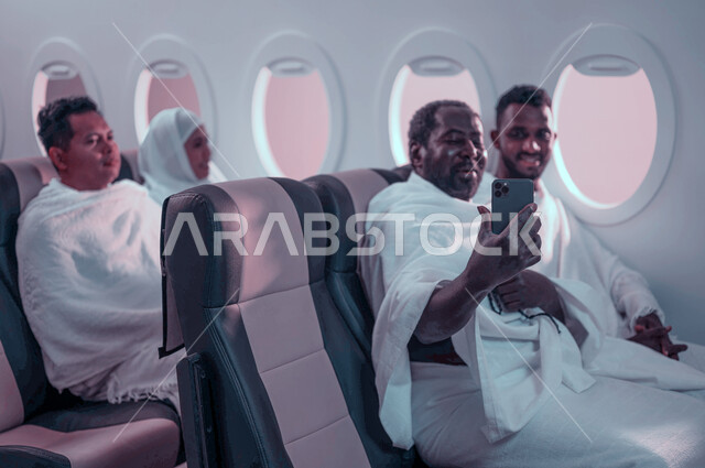 التقاط صور تذكارية من خلال الهاتف المحمول، مجموعة من حجاج الخارج يجلسون في مقصورة الطائرة ويرتدون ملابس الاحرام، بث فيديو مباشر، فضل العمرة في رمضان، السفر الى مكة لآداء مناسك الحج والعمرة، موسم الحج 2023، استقبال الحجاج من خارج السعودية