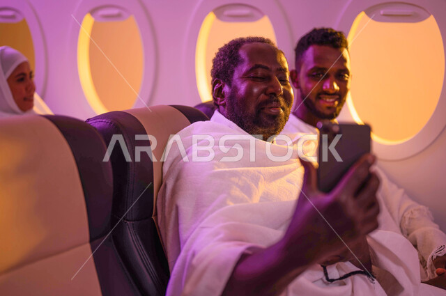 بث فيديو مباشر، التقاط صورة سيلفي تذكارية من خلال الهاتف المحمول، رجلان عربيان من السودان يرتديان ملابس الاحرام ويجلسان في الطائرة، السفر الى مكة لآداء مناسك الحج والعمرة، موسم الحج 2023، آداء العمرة في رمضان، خطوط الطيران السعودي