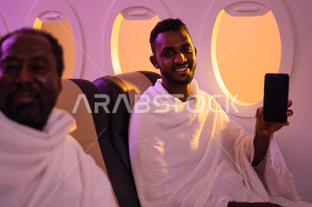 رجلان عربيان من السودان يجلسان في الطائرة ويرتديان ملابس الاحرام، السفر الى مكة لآداء مناسك الحج والعمرة، الهاتف المحمول بشاشة سوداء فارغة، عرض الخدمات والتطبيقات الارشادية لاداء مناسك الحج، موسم الحج 2023، فضل العمرة في رمضان، خطوط الطيران السعودي