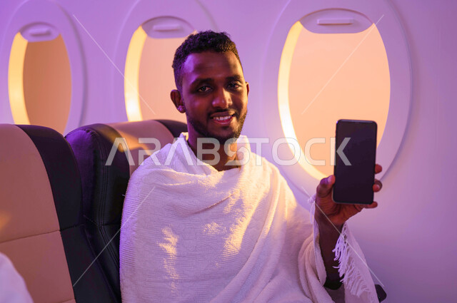 Traveling to Mecca to perform the rituals of Hajj and Umrah, a dark-skinned Sudanese Arab man holding a mobile phone with a blank black screen, Hajj season 2023, online booking for Hajj and Umrah campaigns using mobile phones, performing Umrah in Ramadan,