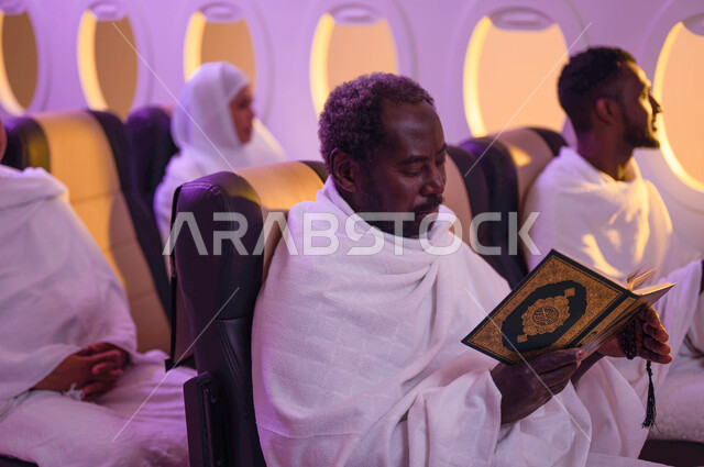 حفظ وتفسير القران الكريم، رجل عربي سوداني اسمر البشرة يقراء القرآن، مجموعة من حجاج الخارج يجلسون في مقصورة الطائرة ويرتدون ملابس الاحرام، السفر الى مكة لآداء مناسك الحج والعمرة، موسم الحج 2023، آداء العمرة في رمضان، استقبال الحجاج من خارج السعودية