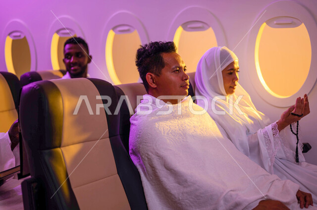 العباده والتقرب الى الله، استقبال الحجاج من خارج السعودية، مجموعة من حجاج الخارج يجلسون في الطائرة ويرتديون ملابس الاحرام، فضل العمرة في رمضان، امرأة اندونيسية مسلمة تمسك السبحة وترفع يدها للأعلى، موسم الحج 2023، السفر الى مكة لآداء مناسك الحج والعمرة