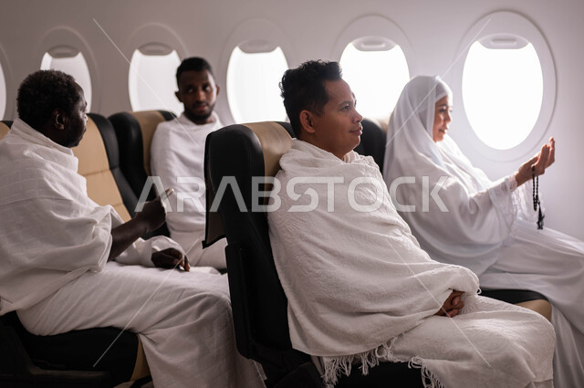 السفر الى مكة لآداء مناسك الحج والعمرة، استقبال الحجاج من خارج السعودية، فضل العمرة في رمضان، موسم الحج 2023، امرأة اندونيسية مسلمة تمسك السبحة وترفع يدها للأعلى، الدعاء والتسبيح والاستغفار، مجموعة من حجاج الخارج يجلسون في الطائرة، ارتداء ملابس الاحرام
