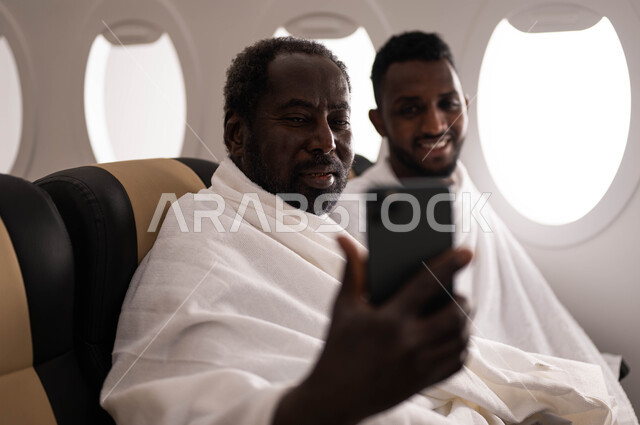 Taking memorial photos via mobile phone, two Arab men from Sudan wearing Ihram clothes and sitting in the seat of the plane, live broadcast video, traveling to Mecca to perform Hajj and Umrah, Hajj season 2023, performing Umrah in Ramadan, Saudi Airlines