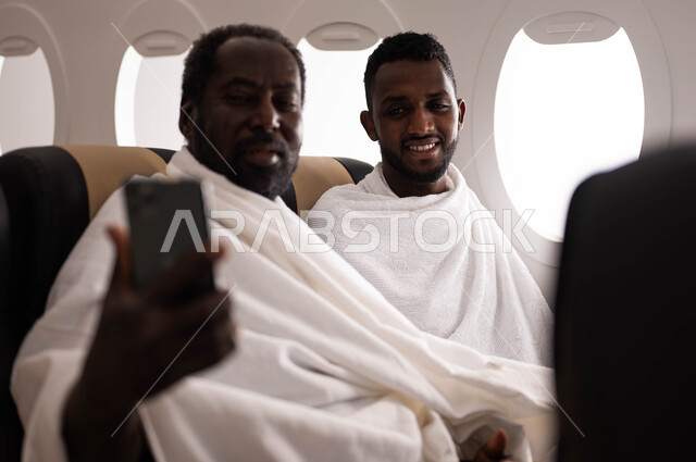 فيديو بث مباشر، السفر الى مكة لآداء مناسك الحج والعمرة، رجلان عربيان من السودان يرتديان ملابس الاحرام ويجلسان في مقعد الطائرة، فضل العمرة في شهر رمضان، التقاط صور تذكارية من خلال الهاتف المحمول، موسم الحج 2023، خطوط الطيران السعودي