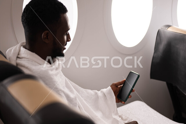Hajj season 2023, online booking for Hajj and Umrah campaigns using mobile phones, wearing Ihram clothes, a dark-skinned Sudanese Muslim youth holding a mobile phone with a black screen and sitting on the plane, traveling to Mecca to perform the rituals o