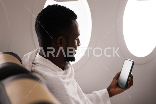 ارتداء ملابس الاحرام، شاب سوداني مسلم اسمر البشرة يمسك الهاتف المحمول بشاشة سوداء ويجلس في الطائرة، استخدام تطبيق الارشادات لاداء مناسك الحج والعمرة، السفر الى مكة لآداء مناسك الحج والعمرة، موسم الحج 2023، آداء العمرة في رمضان، خطوط الطيران السعودي