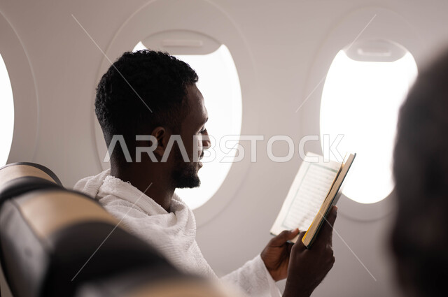 السفر الى مكة لآداء مناسك الحج والعمرة، حفظ وتفسير القران الكريم، شاب سوداني مسلم اسمر البشرة يرتدي ملابس الاحرام ويقوم بقراءة وتلاوة القران، الجلوس في مقعد الطائرة، موسم الحج 2023، رحلة سياحة دينية، فضل العمرة في شهر رمضان، خطوط الطيران السعودي
