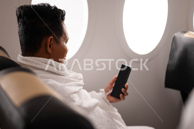 ارتداء ملابس الاحرام، صورة من الخلف لرجل اندونيسي مسلم يمسك هاتف محمول بشاشة سوداء ويجلس في الطائرة، استخدام تطبيق الارشادات لاداء مناسك الحج والعمرة، السفر الى مكة لآداء مناسك الحج والعمرة، موسم الحج 2023، آداء العمرة في رمضان، خطوط الطيران السعودي