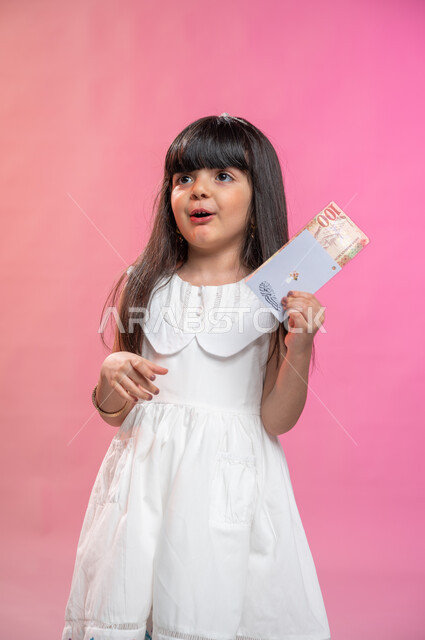 التهنئة بقدوم العيد,   الحصول على العيدية بمناسبة الأعياد السعيدة, ايماءات وجه تدل على السعادة و السرور, بورتريه لطفلة عربية خليجية سعودية تحمل بيدها نقود, أعياد ومناسبات إسلامية, خلفية ملونة.
