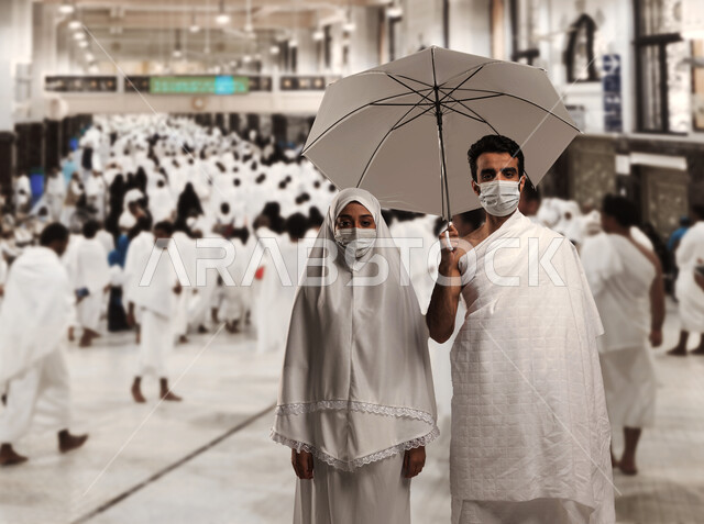 الالتزام بالإجراءات الاحترازية بارتداء الكمامة للوقاية من الأمراض, اداء مناسك الحج والعمره في موسم الحج 2023، زوجان عربيان خليحيان سعوديان يرتديان ثياب الاحرام، الركن الخامس من اركان الاسلام، رجل محرم يحمل بيده شمسية رمادية,  خلفية ممر الصفا و المروة.