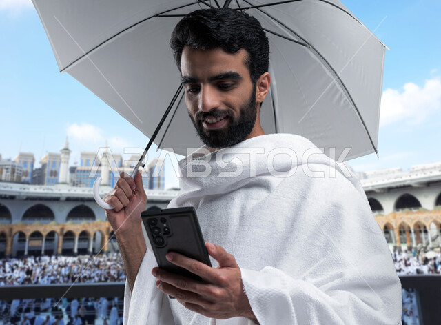 الدليل الالكتروني للحاج و المعتمر, آداء فريضة الحج و العمرة في موسم الحج 2023, بورتريه لرجل مسلم خليجي عربي سعودي يرتدي ملابس الإحرام ويحمل بيده شمسية رمادية ويقوم باستخدام تطبيقات  الهاتف المحمول، خلفية الحرم النبوي الشريف.
