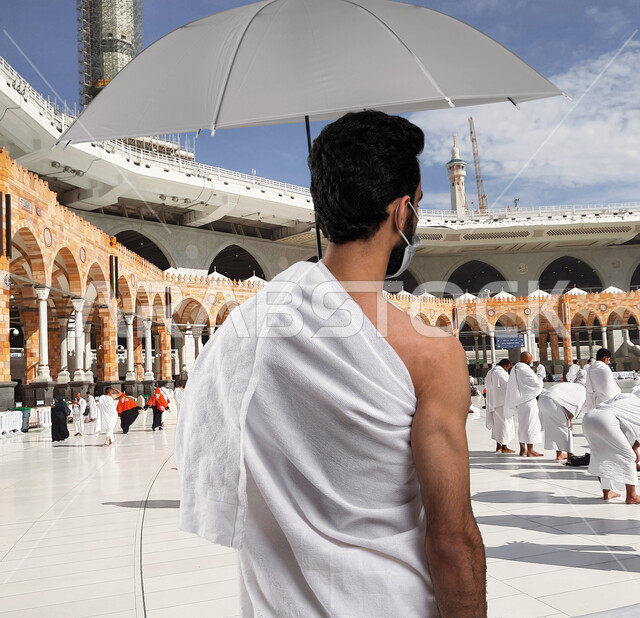 العمرة في شهر رمضان المبارك, شاب عربي خلجي سعودي يرتدي ملابس الإحرام ويحمل بيده مظلة رمادية, زيارة بيت الله الحرام في مكه المكرمه,  فريضة الحج في موسم الحج 2023, ارتداء الكمامة للوقاية من الأمراض, خلفية الحرم النبوي الشريف.