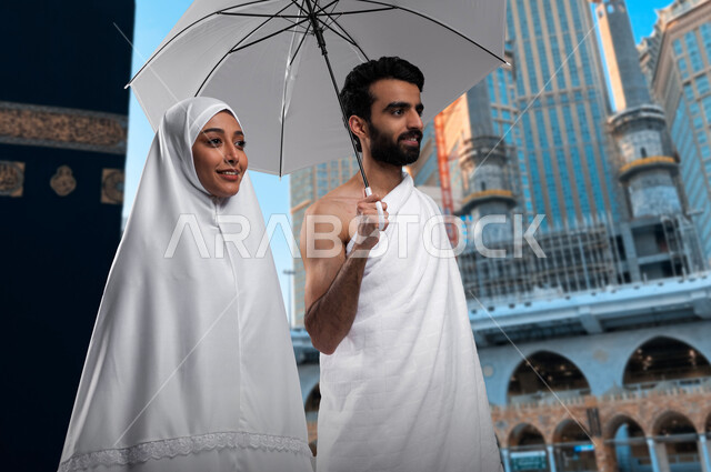 زيارة بيت الله الحرام، أداء مناسك الحج والعمره في موسم الحج 2023، زوجان عربيان خليحيان سعوديان يرتديان ثياب الاحرام ،العمرة في شهر رمضان المبارك شاب يحمل بيده مظلة رمادية، التقرب الى الله بالأعمال الصالحة, خلفية الحرم المكي الشريف.