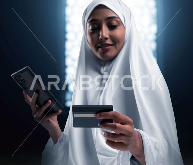 Purchase and payment by credit card, electronic reservation in Hajj and Umrah campaigns, a Saudi Arabian Gulf woman wearing prayer clothes using a mobile phone and a bank card, fast online reservations, an Islamic background.
